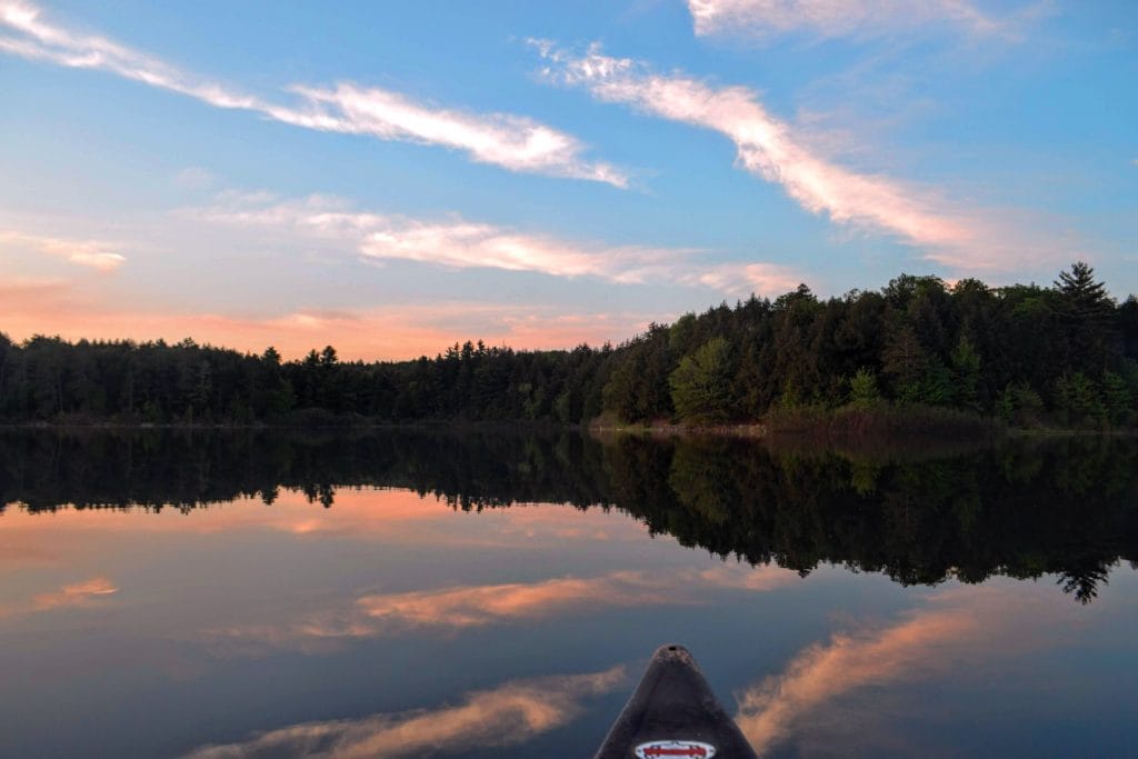 photograph of calm waters on upper peninsula michigan in summer near sunset. evergreen trees line the shore line and water has perfect reflection of the blue and pink sky with whispy clouds, nose of the front of the canoe in the bottom middle. pristine environment