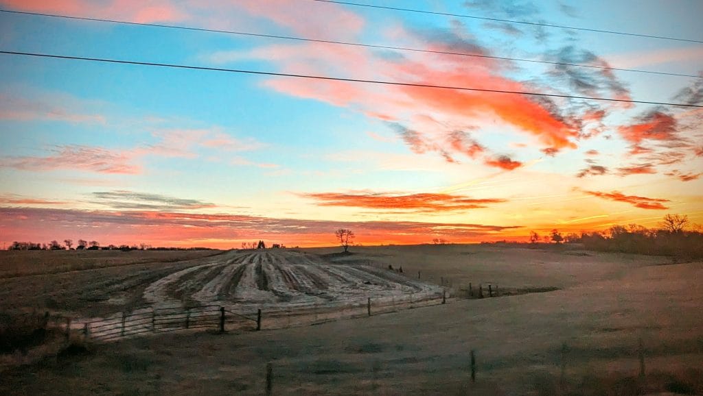 farm in iowa at sunset in winter with some bare fields and some cover crop. horizon balanced in center with blue, pink, and orange skies with some clouds